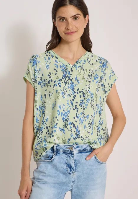 Chemisier à manches courtes avec mélange de textures raw matcha lime Frau in hellgrüner Bluse mit blauem Blumenmuster und Jeans, lächelnd, Hände in den Taschen.
