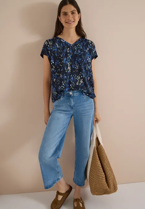 Chemisier à manches courtes avec mélange de textures urban dark blue Frau trägt geblümte Bluse, weite Jeans und hält eine große, braune Tragetasche vor neutralem Hintergrund.