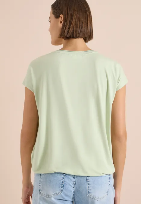 Frau von hinten in hellgrünem T-Shirt und Jeans vor hellem Hintergrund.
