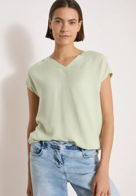 Frau in locker sitzender, hellgrüner V-Neck-Bluse und hellblauen Jeans vor weißem Hintergrund.