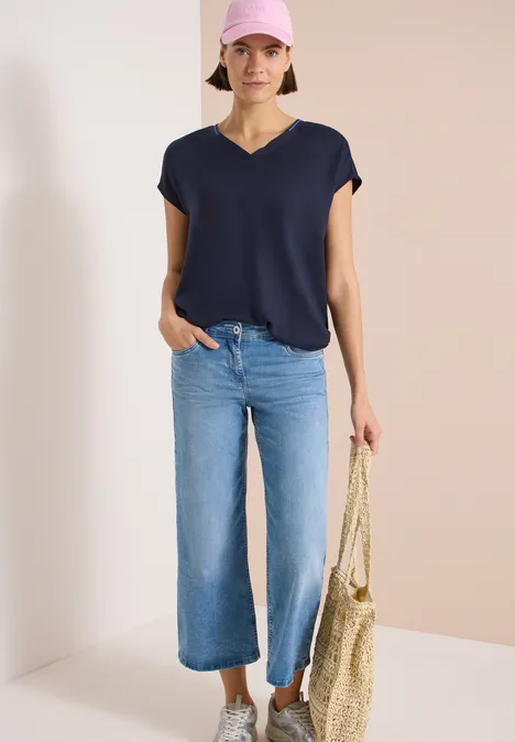 Frau in dunkelblauem V-Shirt und weit geschnittener blauer Jeans mit Strohtasche vor schlichtem Hintergrund.