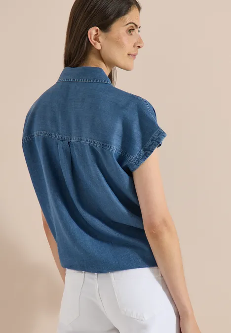 Chemisier à épaules tombantes, effet denim mid blue wash Une femme porte un chemisier en jean à manches courtes et un pantalon blanc, vue de dos sur fond clair.