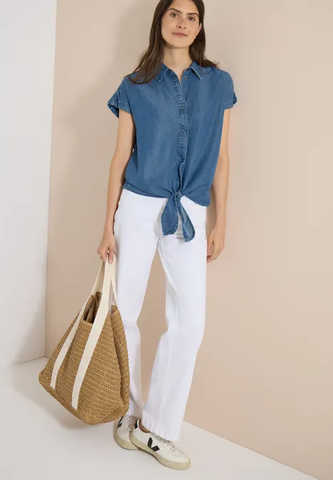 Chemisier à épaules tombantes, effet denim mid blue wash Femme en chemise bleue nouée, pantalon blanc et sac en paille sur fond beige.