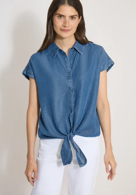 Chemisier à épaules tombantes, effet denim mid blue wash Haut en jean sans manches noué à l'avant, porté avec un pantalon blanc sur fond neutre.