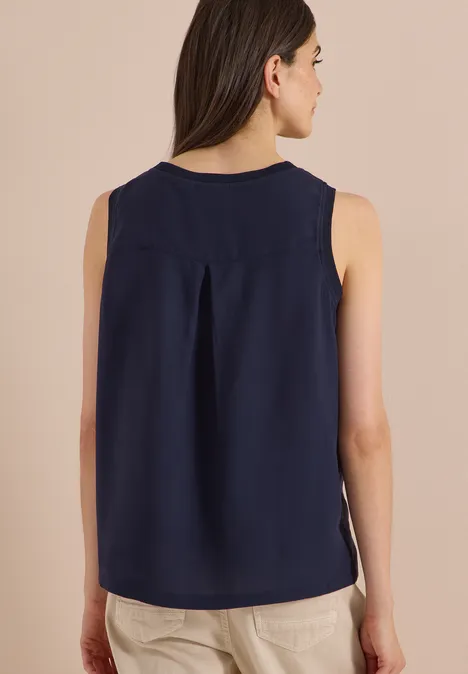 Rückenansicht einer Person in ärmelloser, dunkelblauer Bluse mit rückseitlicher Mittelnaht und beiger Hose.