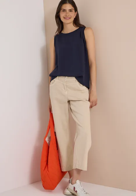 Frau in dunkelblauem Top und weiten, beigen Culotte-Hosen hält eine orange Tasche.