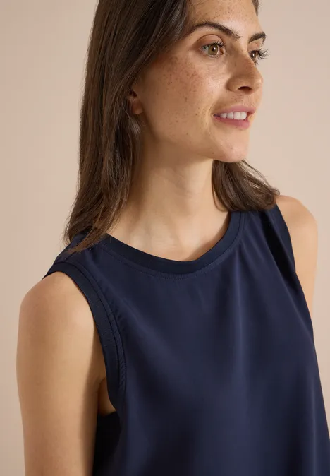 Eine Frau mit dunklem ärmellosem Top, leichtem Make-up und braunen Haaren, blickt seitlich und lächelt.
