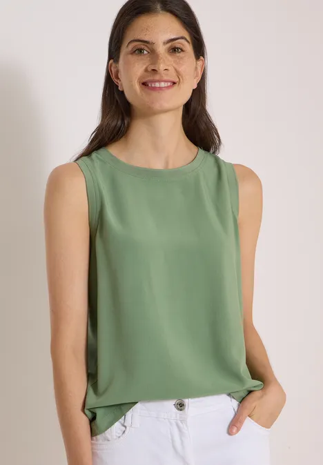 Eine lächelnde Frau in ärmellosem grünem Top und Jeans vor hellem Hintergrund.