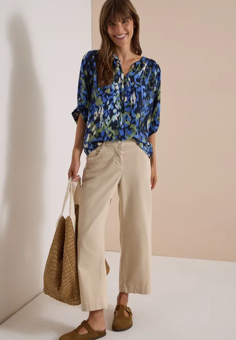 Frau in blau-gemusterter Bluse, beigen weiten 7/8-Hosen, braunen Mokassins und Strohtasche, lächelnd.