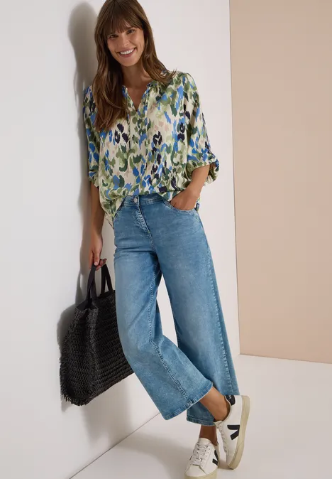 Eine lächelnde Frau trägt eine gemusterte Bluse, gerade geschnittene Jeans, weiße Sneaker und hält eine Tasche.
