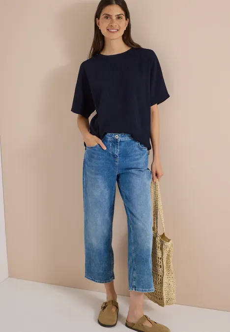 Chemisier à épaules tombantes, couleur unie urban dark blue Eine Frau trägt ein dunkelblaues T-Shirt, weite blaue Jeans und Sandalen, hält eine geflochtene Stroh-Tasche.