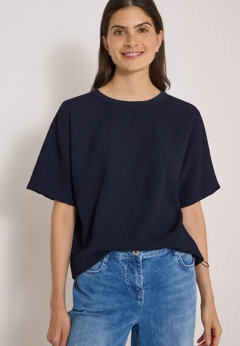 Chemisier à épaules tombantes, couleur unie urban dark blue Eine lächelnde Frau in dunkelblauem T-Shirt und Jeans vor weißem Hintergrund.