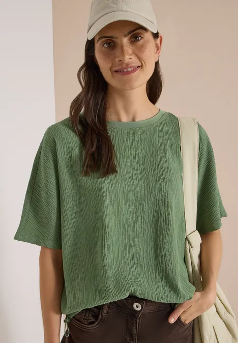 Lächelnde Frau in grünem, strukturiertem T-Shirt, beige Cap und Canvas-Tasche vor neutralem Hintergrund.