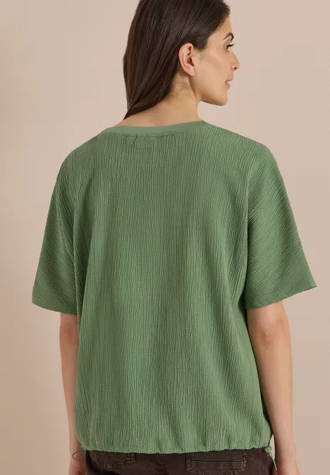 Frau von hinten in locker sitzendem, kurzärmeligem, grünem, leicht gekräuseltem Shirt vor beigem Hintergrund.