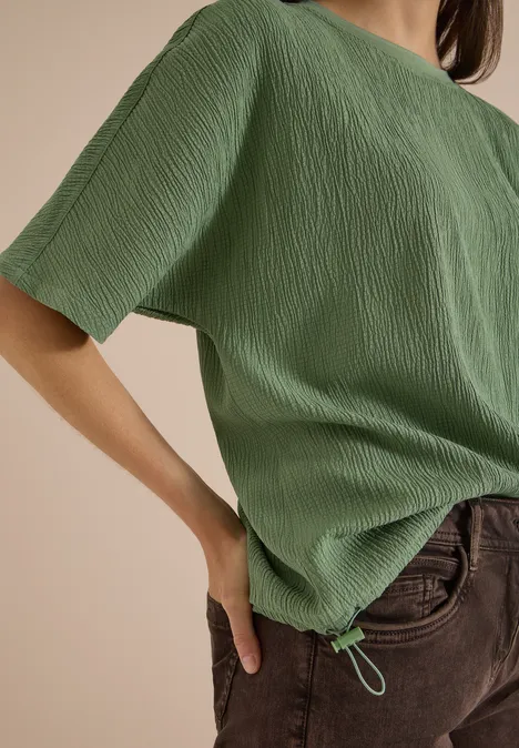 Eine Person trägt ein grünes, strukturiertes Kurzarmshirt mit Kordelzugsaum und braune Hosen