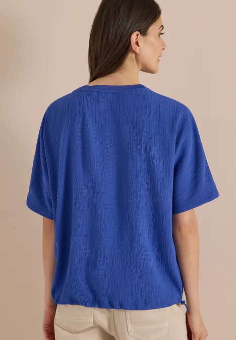 Chemisier à épaules tombantes, couleur unie deep river blue Eine Frau mit dunklen Haaren trägt eine blaue Bluse und helle Hose, von hinten vor beigem Hintergrund.