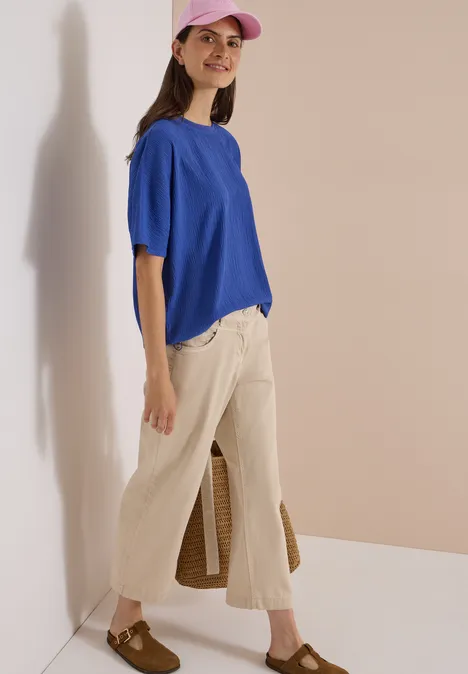 Chemisier à épaules tombantes, couleur unie deep river blue Eine Frau mit rosa Baseballcap, blauem T-Shirt, beigen Culottes und braunen Loafers vor hellem Hintergrund.
