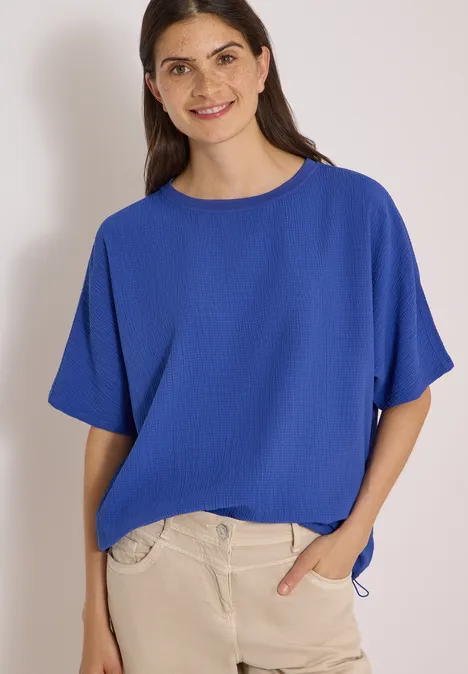 Chemisier à épaules tombantes, couleur unie deep river blue Frau in lockerem blauen T-Shirt mit kurzen Ärmeln und heller Hose, lächelnd vor hellem Hintergrund.