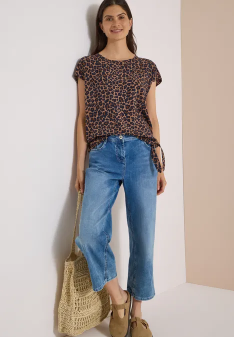 Chemisier à épaules tombantes avec nœud creamy mocha Eine Frau trägt ein gepunktetes Shirt, weite blaue Jeans, Keilsandalen und hält eine geflochtene Tasche.