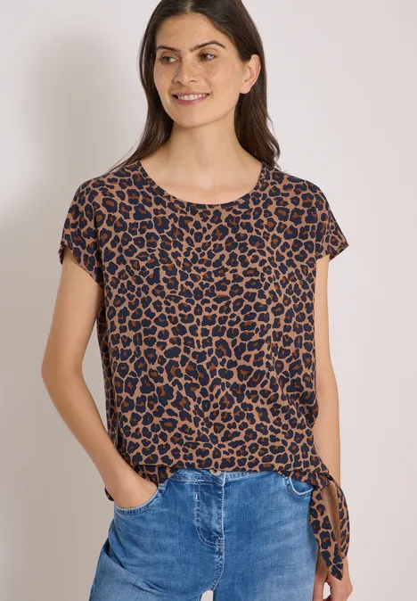 Chemisier à épaules tombantes avec nœud creamy mocha Frau in braun-schwarzem Leoparden-T-Shirt mit seitlicher Schleife und blauen Jeans.