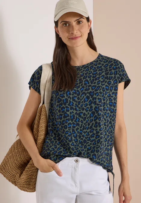 Chemisier à épaules tombantes avec nœud dried olive green Frau in blau-grünem Leo-Shirt, weißer Hose und Strohtasche, mit Cap, lässig lächelnd
