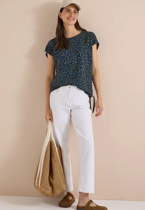 Chemisier à épaules tombantes avec nœud dried olive green Frau mit gemusterter Bluse, weißer Leinenhose, Strohtasche, Cap und braunen Loafers.