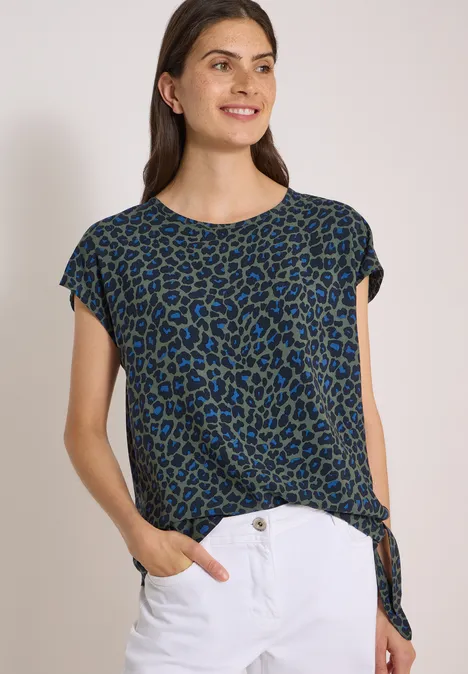 Chemisier à épaules tombantes avec nœud dried olive green Frau trägt blau-grünes Leo-Shirt mit seitlicher Knoten-Optik und weiße Hose, lächelnd.
