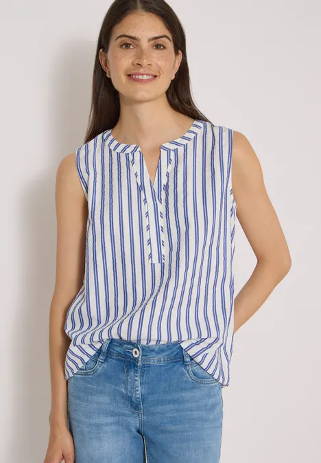 Eine lächelnde Frau in blau-weißer, ärmelloser Bluse mit Knopfleiste, kombiniert mit blauen Jeans.