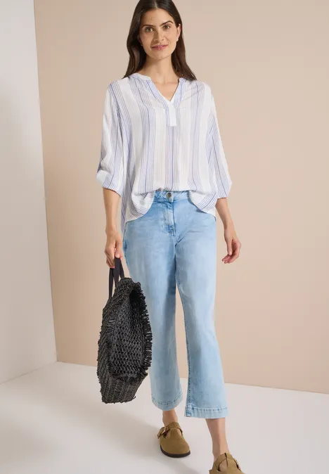 Frau trägt hellblau-weiß gestreifte Bluse, lockere Jeans, beige Slipper und hält einen schwarzen, gewebten Shopper.