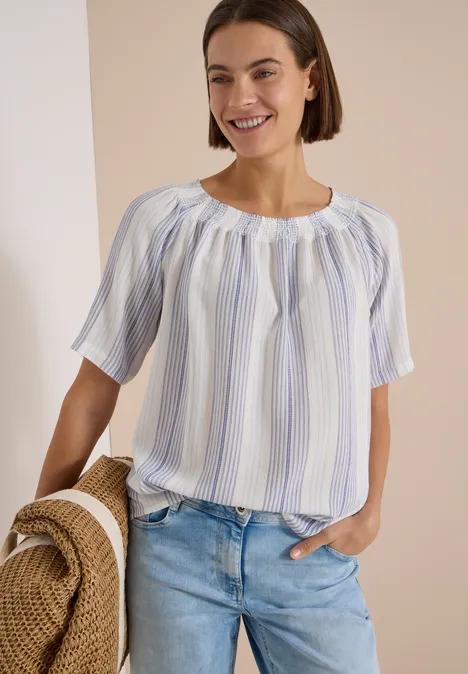 Carmen-Bluse mit Streifenmuster deep river blue Frau trägt helle, gestreifte Bluse und Jeans, hält einen Strohhut und lächelt.