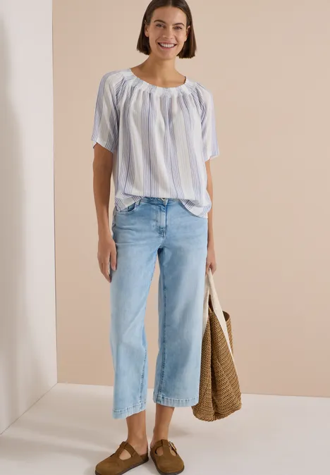 Carmen-Bluse mit Streifenmuster deep river blue Eine lächelnde Frau in gestreiftem, lockerem Oberteil, ausgestellten, hellen Jeans, braunen Loafers und Korbtasche.