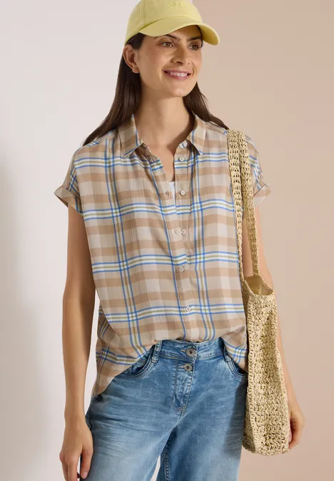Eine junge Frau trägt eine beige-karierte Bluse, blaue Jeans, eine Strohtasche und eine gelbe Mütze und lächelt.