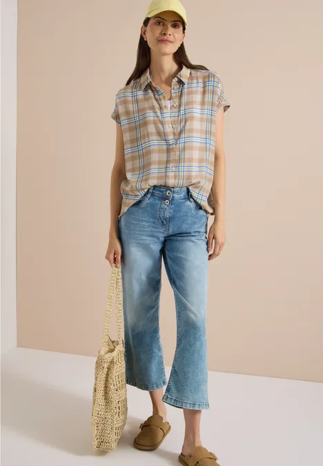 Junge Frau in karierter Bluse, weiter Jeans, Cap, Strohtasche und Pantoletten vor neutralem Hintergrund.