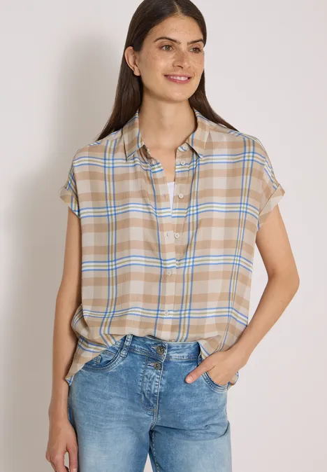 Frau in beige-kariertem Kurzarmhemd und blauen Jeans, lächelnd und leger posierend.