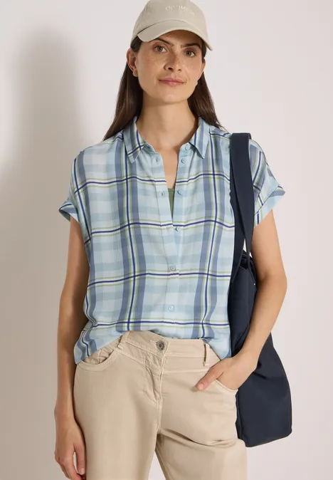Femme en chemise bleue à carreaux et casquette, pantalon beige et sac bleu, posant de façon décontractée.
