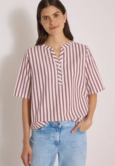 Chemisier à manches courtes, à col fendu et rayures russet red Frau in gestreiftem rot-weißem Blusenshirt und blauen Jeans vor hellem Hintergrund.