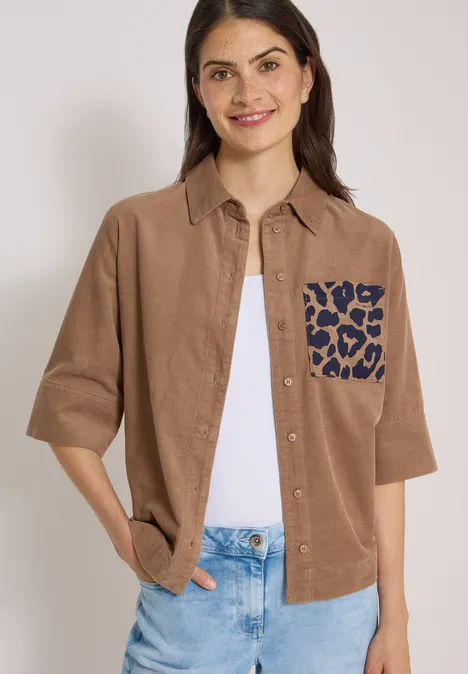 Overhemd met korte mouwen van corduroy met borstzak creamy mocha Lächelnde Frau in braunem Cordhemd mit blau gefleckter Leoparden-Tasche, weißem Top und Jeans.
