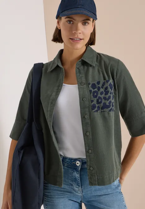 Overhemd met korte mouwen van corduroy met borstzak dried olive green Frau trägt grüne Bluse mit Leopardenmuster-Tasche, weißem Top, Jeans, dunkelblauer Cap und Tasche.