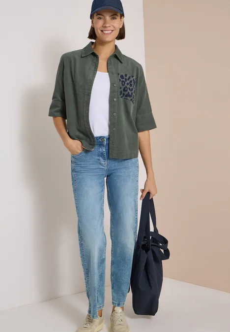 Overhemd met korte mouwen van corduroy met borstzak dried olive green Frau in Jeans, weißem T-Shirt, grüner Bluse, blauer Mütze trägt eine große blaue Tasche.