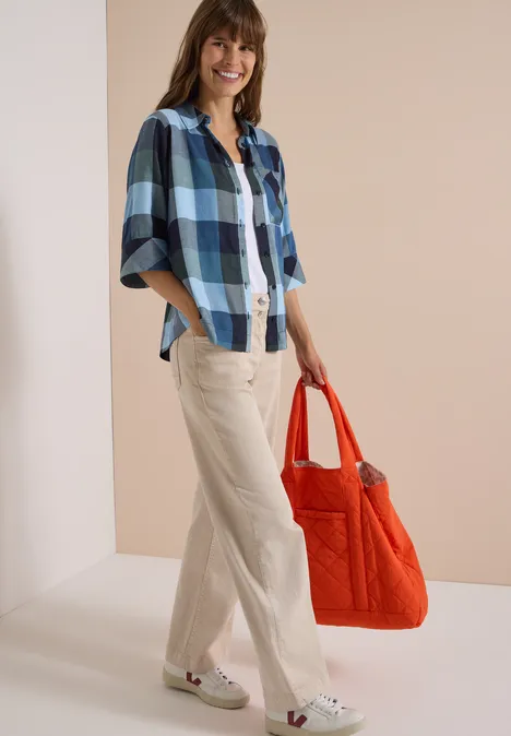 Lächelnde Frau in blau-weiß kariertem Hemd, heller Hose und orangefarbener Tasche.