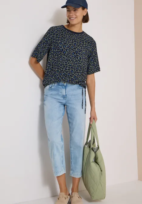Blouse met korte mouwen en luipaardprint dried olive green Junge Frau in schwarzem, gemustertem T-Shirt, hellen Jeans, Cap und grüner Stepp-Tasche.