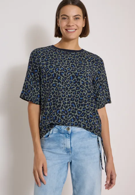 Blouse met korte mouwen en luipaardprint dried olive green Eine Frau lächelt trägt ein blau-schwarz gemustertes T-Shirt mit Knoten am Saum und helle Jeans vor weißem Hintergrund