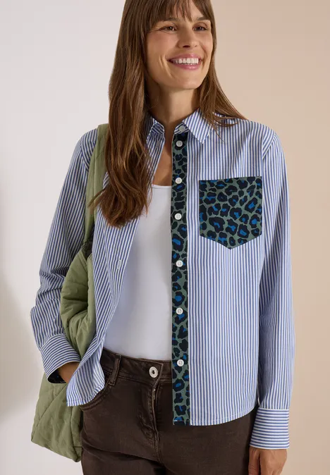 Hemdbluse mit Streifen und Leo-Brusttasche blouse blue Frau lächelt, trägt blau-weiß gestreiftes Hemd mit Blumenbesatz, grüne Steppjacke am Arm und braune Hose.