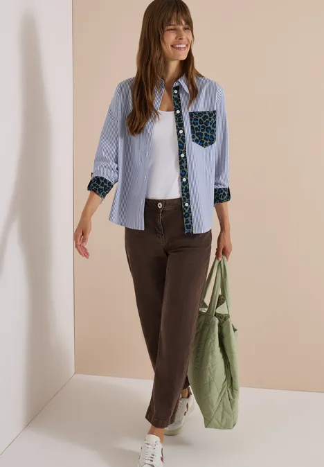 Hemdbluse mit Streifen und Leo-Brusttasche blouse blue Frau trägt hellblaues Hemd mit gemusterten Details, braune Hose und grünen Beutel, lächelt.