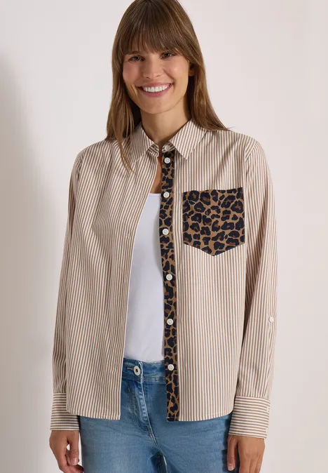 Hemdbluse mit Streifen und Leo-Brusttasche creamy mocha Frau trägt beige Hemd mit Leopardenmuster an Brusttasche und Knopfleiste, darunter weißes Top und Jeans.