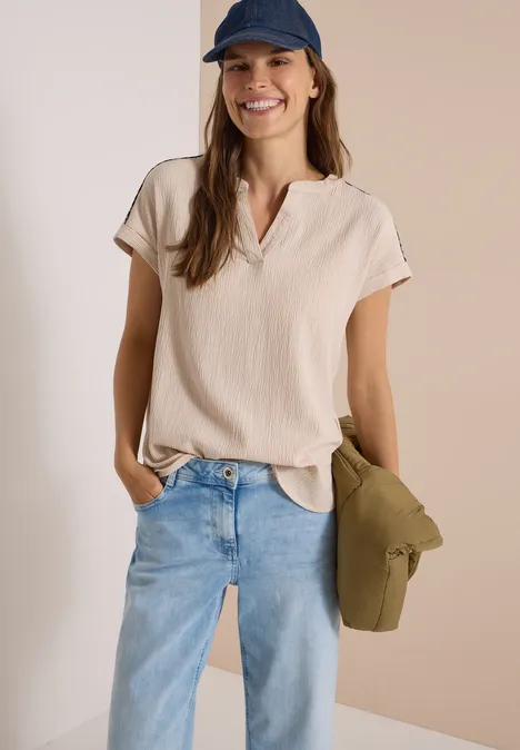 Frau mit blauer Cap, hellbeigefarbenem Shirt, hellblauen Jeans und einer grünen Tasche, lächelt lässig.