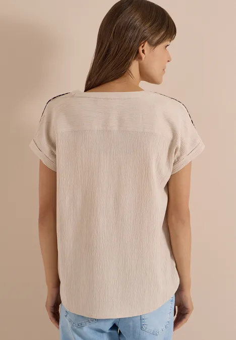 Eine Frau von hinten in einer hellbeigen, kurzärmeligen Bluse und blauen Jeans vor neutralem Hintergrund.