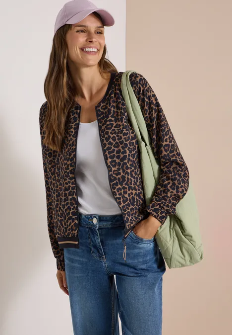 Blouson mit Zipper und Leo-Muster creamy mocha Frau mit Baseballcap, Leoparden-Bomberjacke, weißem Top, Jeans und grünem Shopper vor zweifarbigem Hintergrund.