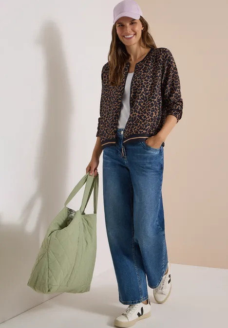 Blouson mit Zipper und Leo-Muster creamy mocha Frau in Leopardenjacke, weißem Top und blauer Jeans trägt grünen Shopper und rosa Baseballcap.