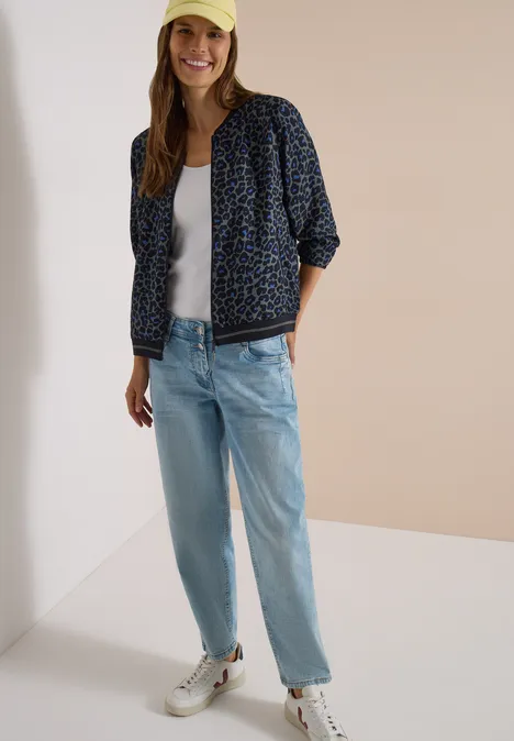 Femme en casquette jaune, veste bomber à motifs, top blanc, jean clair et baskets blanches sur fond neutre.